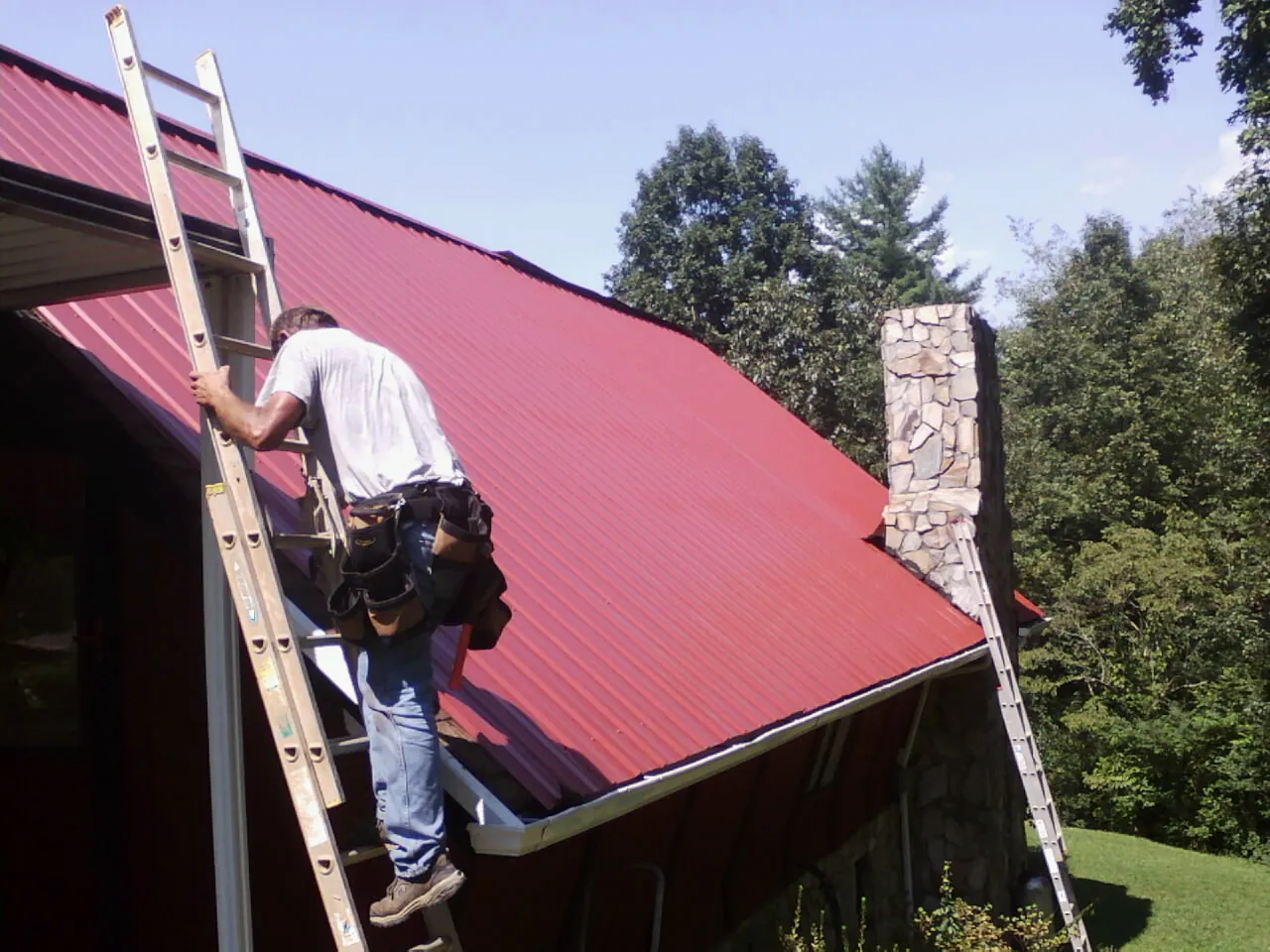 Roofer on a ladder beside a red metal roof and stone chimney — Soffit & Fascia Repair by Roofing Brothers in Mad River